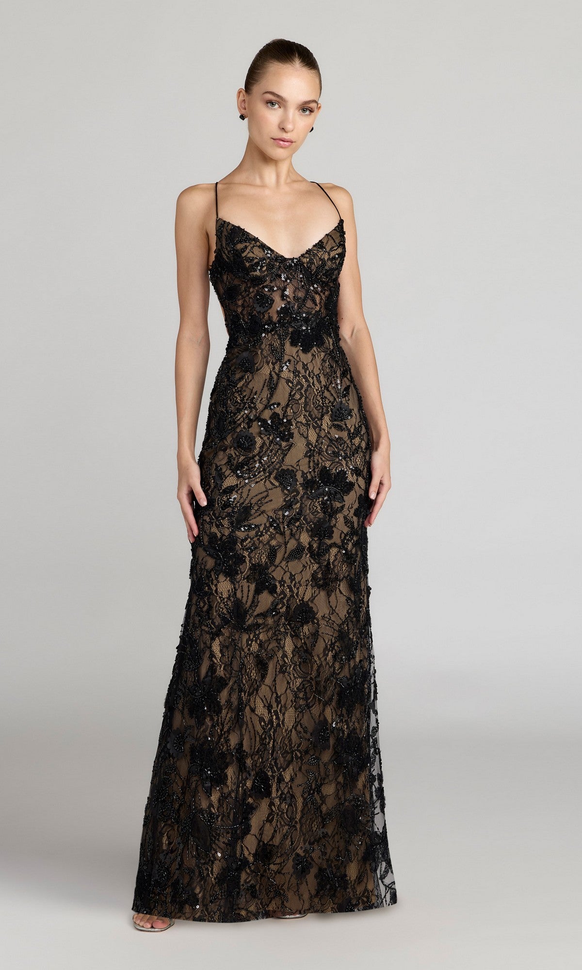 A woman models the Mac Duggal 68892 long formal dress in Black Nude, featuring a floor-length, sleeveless design with an open back, thin straps, and black beaded lace embroidery against a plain light gray background.